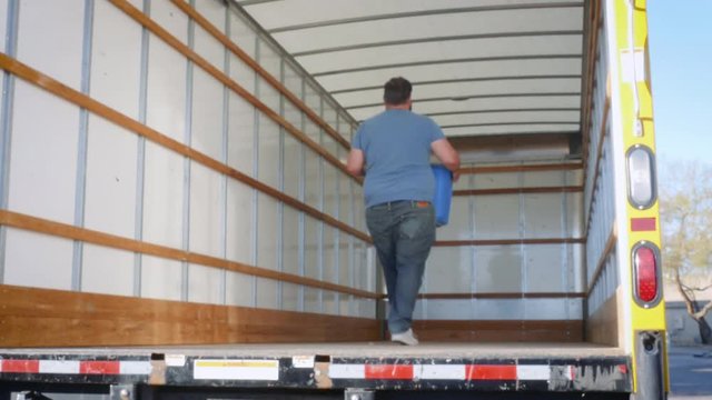Man loads a moving truck with a blue plastic box in 4k. The young man walks up the metal ramp with a bin of belongings preparing to relocate to a new city. Candid shot represents relocating for work.