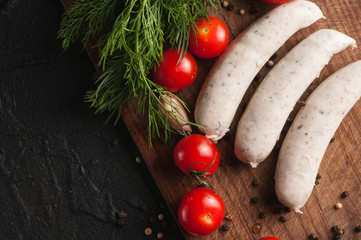 Three raw sausages on a wooden brown board on a black background with cherry tomatoes, parsley and dill, pepper and garlic.