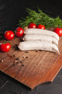 Three raw sausages on a wooden brown board on a black background with cherry tomatoes, parsley and dill, pepper and garlic.