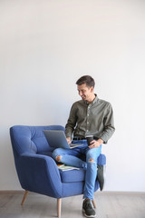 Young blogger with laptop sitting on armchair against light wall