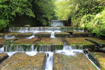 Fototapeta na wymiar Summer scenery of beautiful waterfalls cascading down a stream in a shady jungle in Taiwan ~ Cool refreshing cascades hidden in a mysterious forest of lush greenery
