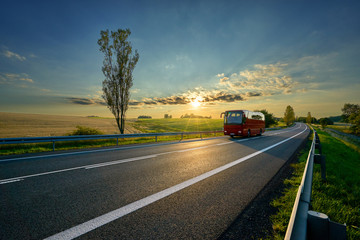 Red bus traveling on asphalt road around farm fields in rural landscape at sunset