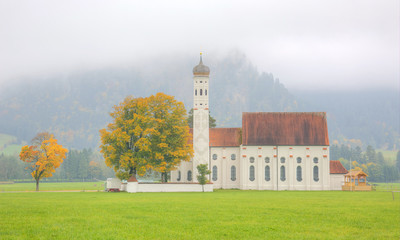 Fall scenery of Bavarian countryside in Schwangau, Germany, Europe, with view of majestic St. Coloman Church and beautiful trees standing in the green meadow on a foggy autumn morning