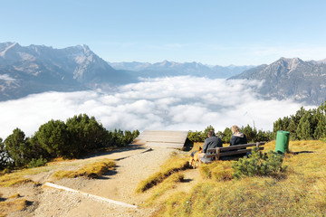 Naklejka premium Tourists sitting on wooden bench and enjoying the beautiful mountain panorama of Zugspitze from top of Mount Wank in Garmisch Partenkirchen, Bavaria, Germany