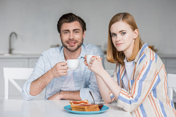 smiling young couple with coffee cups having breakfast at table in kitchen