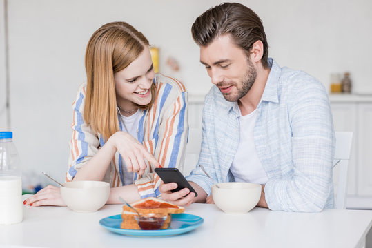 Smiling Young Couple Using Smartphone And Sitting At Table With Breakfast