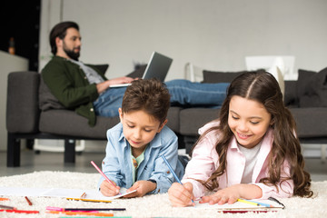 cute smiling kids lying on carpet and drawing with colored pencils while father using laptop on sofa behind