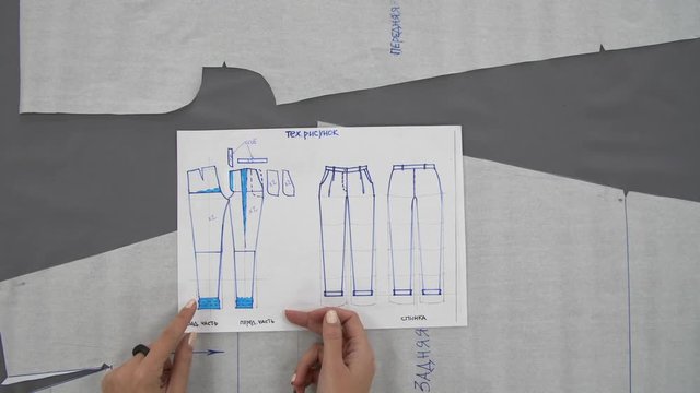 Female hands show something on a pattern for sewing trousers made of gray fabric