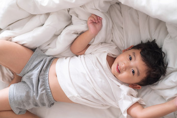 3 years old little cute Asian boy at home on the bed, kid lying playing and smiling on white bed with pillow and blanket, top view with copy space.