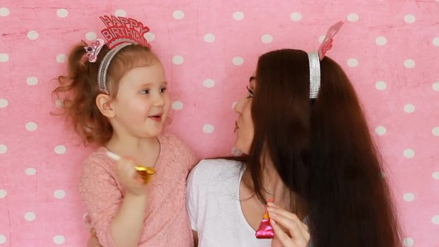 Happy birthday. A woman and her child smiles, have fun, laugh and celebrate, blowing party horns. Close-up portrait Mother and daughter on pink background.