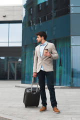 Go forward to succeed! Attractive handsome young newcomer pulling bag on city street near the airport building, drinking coffee-to-go and looking back.