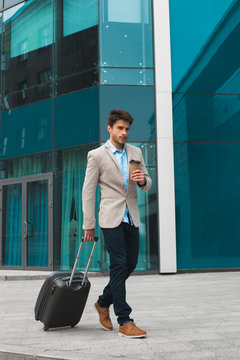 Success is often achieved! Full length vertical shot of young handsome male business traveler pulling baggage on city street, while drinking coffee.
