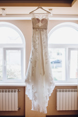 beautiful lace wedding dress on white wooden hanger in luxury room at window in the morning sunlight. space for text. rich wedding concept