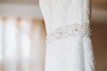 Close-up of fashionable white wedding dress on a hanger in hotel room, morning wedding preparation concept