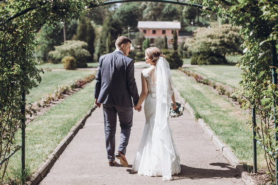 Beautiful Gorgeous Bride And Groom Posing In Sunny Spring Park Among Green Leaves. Happy Wedding Couple Walking In Green Garden. Happy Marriage Moments. Elopement Concept