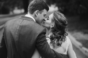 Romantic newlyweds couple hugging and kissing outdoors in summer park, stylish bride in wedding dress on a walk with handsome groom