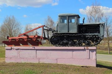Fototapeta premium Grey Soviet crawler tractor with plow on pedestal in spring on blue sky background with clouds