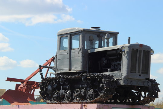 Grey Soviet crawler tractor with plow on pedestal in spring on blue sky background with clouds