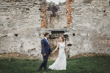 Newlywed couple posing near old castle wall, fairytale wedding at ancient castle outdoors, bride and groom hugging near fortress tower, wedding moment