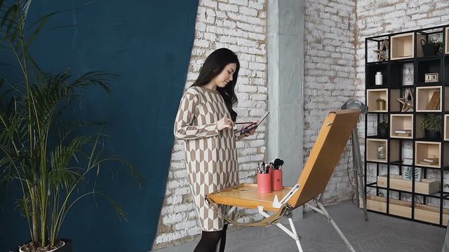 The artist on makeup and beauty creates a beautiful face makeup chart using watercolor paper, shadows, lipsticks and blushes. Young woman in dress, in a bright studio draws using board of easel