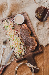 Top view grilled ribeye beef steak with stew cabbage and wineglass of red wine, herbs and spices over brown sackcloth on wooden table,brown brick wall background.