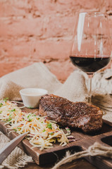 Grilled ribeye beef steak with stew cabbage and wineglass of red wine, herbs and spices over brown sackcloth on wooden table,brown brick wall background.