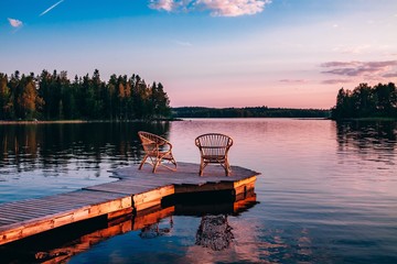 Two wooden chairs on a wood pier overlooking a lake at sunset - Powered by Adobe