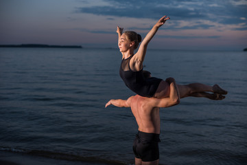 Fototapeta premium Young guy and girl perform beautiful dance elements on the beach on the sand in bathing suits. Holding hands, performs support. Ballet.