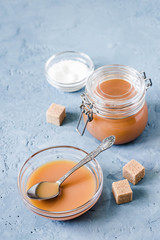Homemade salted caramel sauce in glass jar on light blue concrete background. Selective focus, space for text.