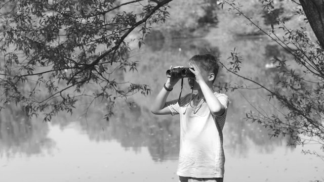 Black and white video of cute funny little kid looks through old vintage binoculars at distance while standing at scenic river bank at fresh foliage of spring tree background.