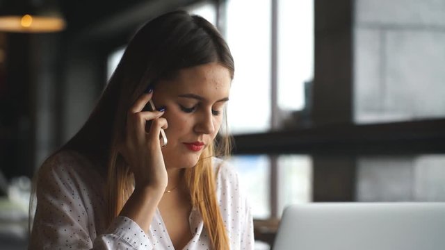 Beautiful young woman working with laptop and talking on smartphone talking to customer sitting near window