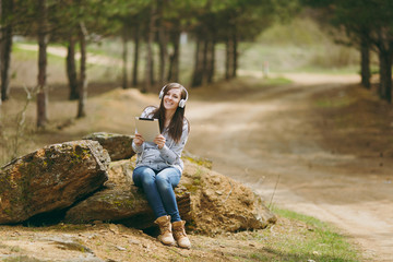 Naklejka na ściany i meble Young smiling beautiful woman in casual clothes with headphones sitting on stone using tablet pc computer in city park or forest on green blurred background. Student lifestyle, leisure concept.