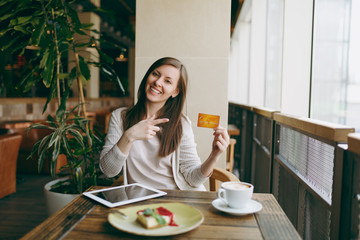 Woman sitting near big window in coffee shop at table with credit card, cup of coffee cake relaxing in restaurant during free time. Female working on tablet pc computer rest in cafe. Lifestyle concept