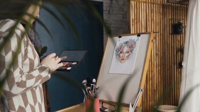 The artist on makeup and beauty creates a beautiful face makeup chart using watercolor paper, shadows, lipsticks and blushes using an easel at studio. Young woman in a bright studio draws a face chart