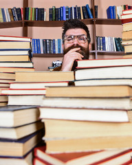 Man on thoughtful face between piles of books in library, bookshelves on background. Scientific research concept. Teacher or student with beard wears eyeglasses, sits at table with books, defocused.