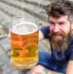 Guy raising up glass with draught beer. Hipster on smiling face drinks beer outdoor. Cheers concept. Man with beard and mustache holds glass with beer while sits on stone stairs, defocused.