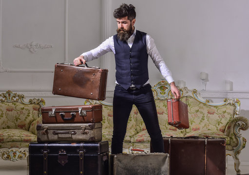 Macho elegant on thoughtful face standing near pile of vintage suitcase. Man, traveller with beard and mustache packing luggage before trip, luxury interior background. Luggage and travelling concept.