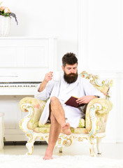 Concentrated guy reading a book while enjoying his morning coffee. Bearded man sitting in armchair with his legs crossed. Serene morning