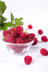 Red fresh raspberries in a glass bowl with green leaves on white background.