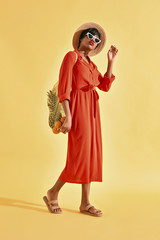 Beautiful young black woman in white sunglasses and hat, dressed in orange red dress , straw shoes, with yellow net bag. Yellow background