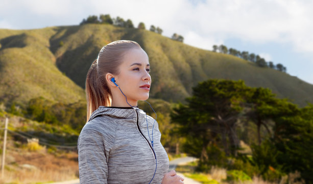 fitness, sport and technology concept - happy woman running and listening to music in earphones over big sur hills background in california