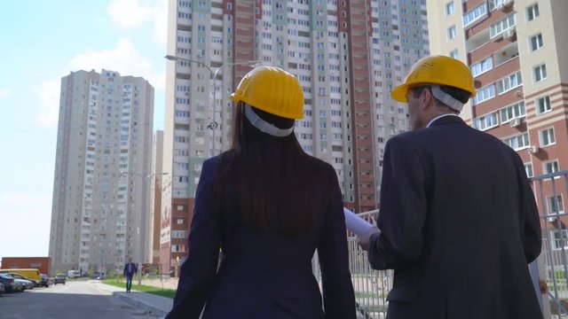 30 years old young woman and middle-aged man go on construction. business woman engineer in yellow helmet holds in hand a planet with papers and smiling. man on the background of the building under