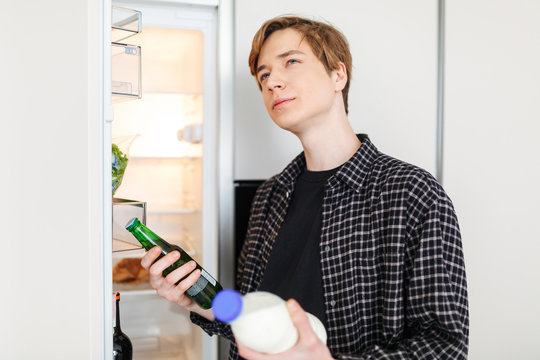 Portrait Of Young Man Standing Near Fridge With Beer And Milk In Hands And Deciding What To Drink In Kitchen At Home Isolated