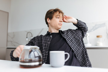 Portrait of young man sitting and pouring coffee while tiredly looking aside in kitchen at home isolated