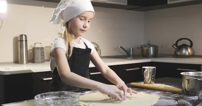 Sweet girl in an apron and chef cap cut out the shape on the cookie dough.