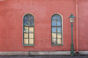 Fototapeta na wymiar House Brick Wall Facade with Two Classic Windows and Street Lantern. Architecture Building Exterior with Red Brick Wall Background and Classic Windows. Outdoor View of Old Architectural House Front.