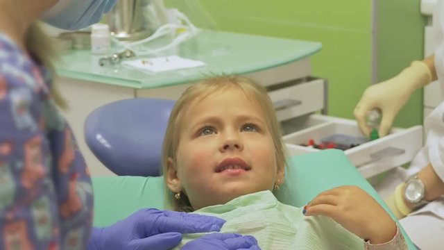 Child with a mother at a dentist's reception. The girl lies in the chair, behind her mother. The doctor works with an assistant. Procedure for drilling a tooth. Setting a temporary seal. Using the x