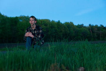 Young man in checkered shirt sitting in grass near country road at dusk.