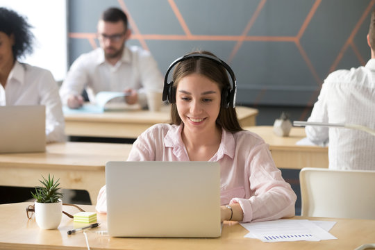 Millennial smiling businesswoman wearing headphones working on laptop in coworking, young manager in headset consulting client online or listening to music while using computer in shared office