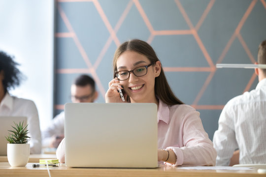 Millennial smiling businesswoman using laptop talking on phone at workplace, young sales manager making call consulting corporate client customer selling by mobile working on computer in coworking